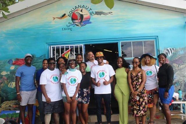 A large group of young people in Earth Day ’24 “Make a Change” T-shirts outside a dive shop in Grenada, one holding the flag of Grenada.