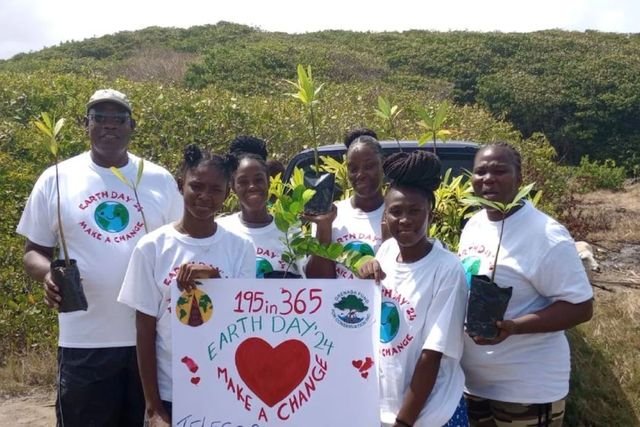 Six people in Earth Day ’24 T-shirts hold potted mangrove seedlings and a “195in365 Earth Day ’24 Make a Change” sign in front of coastal scrubland.
