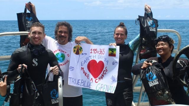 Divers in wetsuits on a boat hold up mesh bags of rubbish and a hand-painted “195in365 Earth Day ’24 Make a Change, TrueBlue Grenada” banner, with open sea behind them.