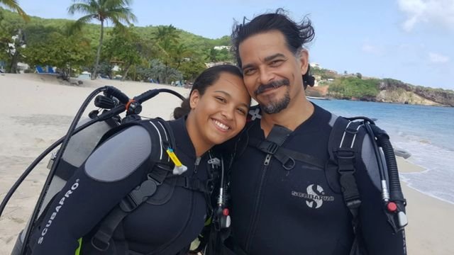 A smiling young woman and a man in wetsuits and scuba gear pose cheek to cheek on a sunny beach, with palm trees and the sea behind them.