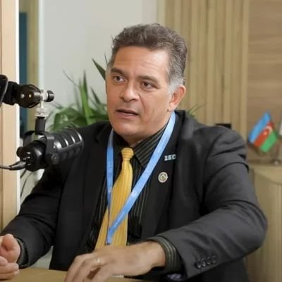 A man in a suit and a conference lanyard speaks into a studio microphone, with a flag on the table behind him.