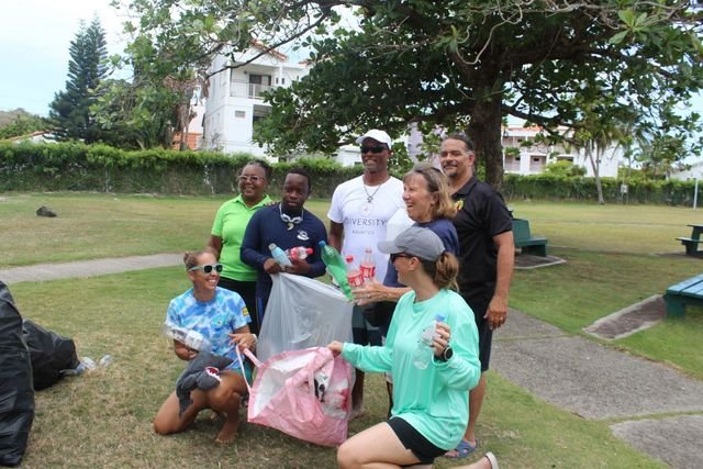 Seven volunteers gather under a large tree in a park, holding plastic bottles and bags of collected rubbish and laughing together.