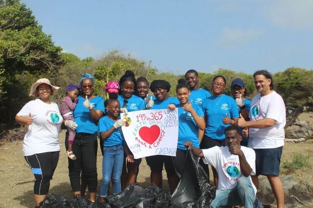 A group of volunteers, most in blue T-shirts and work gloves, give thumbs-up around a “195in365 Earth Day ’24 Make a Change, Grenada” sign beside full rubbish bags.