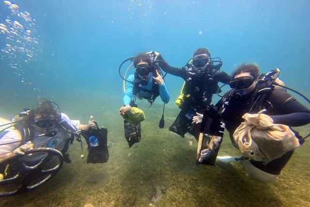 Four scuba divers underwater over a sandy seabed, each holding a mesh bag of collected litter; one gives a thumbs-up.
