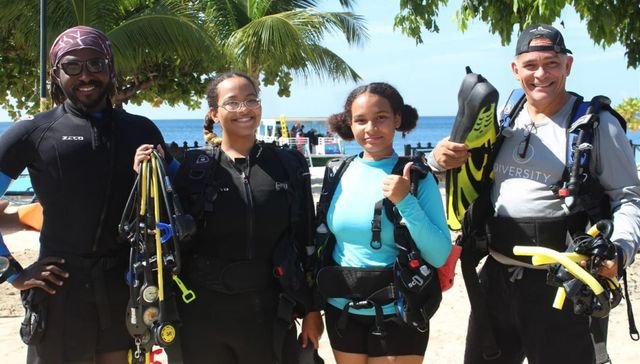 Four divers of different ages stand on a beach holding fins and dive equipment, smiling, with palm trees and a dive boat behind them.