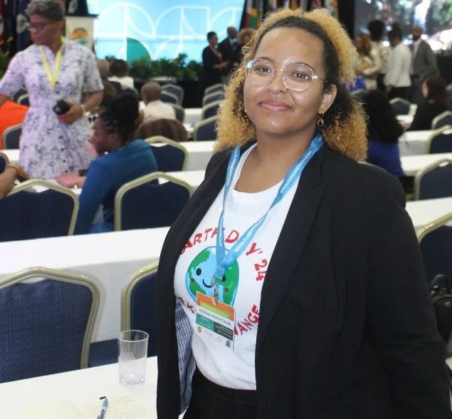 A young woman with glasses and a conference lanyard, wearing an Earth Day ’24 T-shirt under a blazer, stands in a busy conference hall.