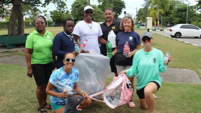 Seven volunteers pose on the grass in a park, two kneeling in front, holding bags full of collected plastic bottles.